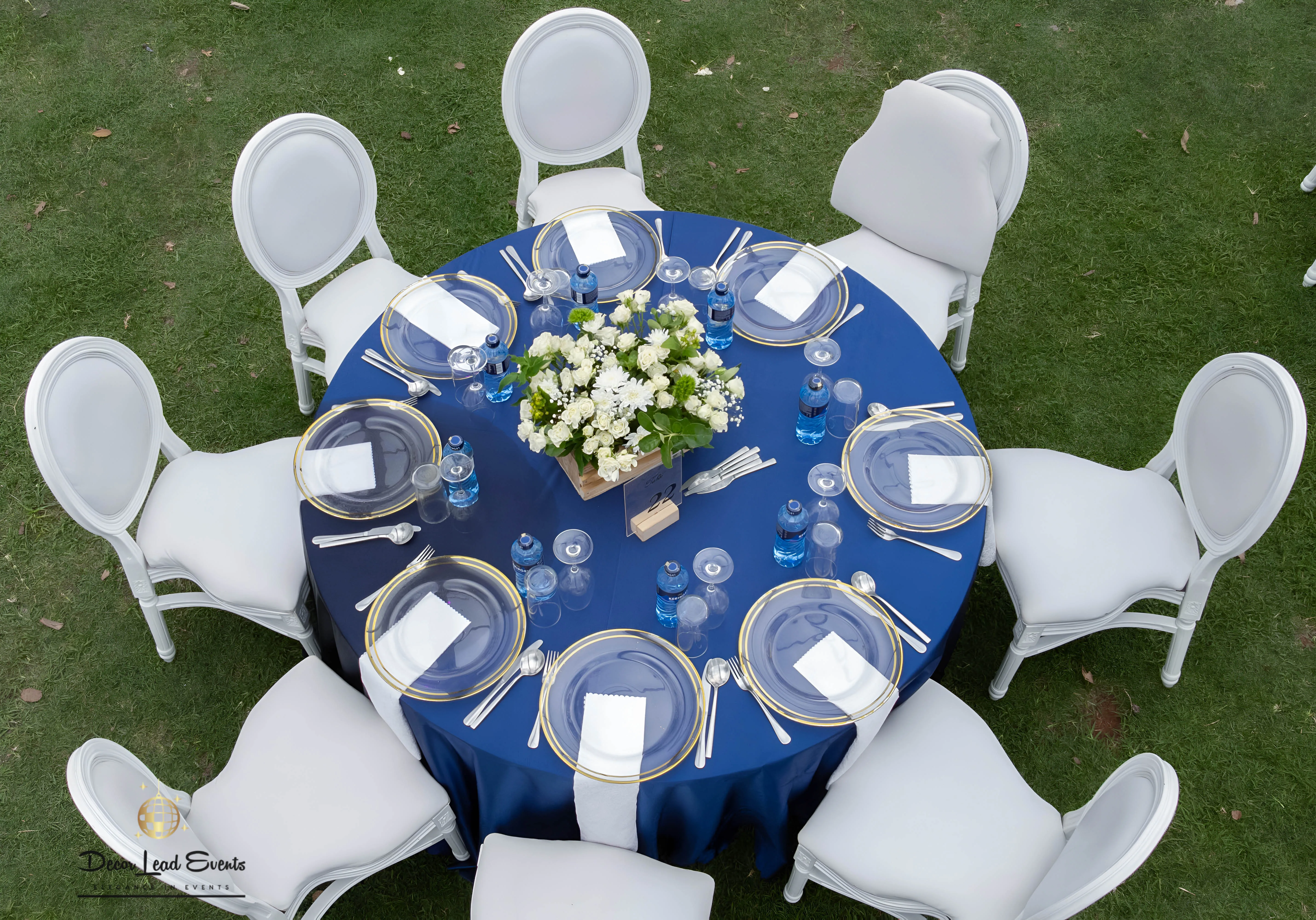 Aerial view of a round table set up with a navy blue tablecloth, gold-rimmed clear glass chargers, white napkins, wine glasses, and a white floral centerpiece.