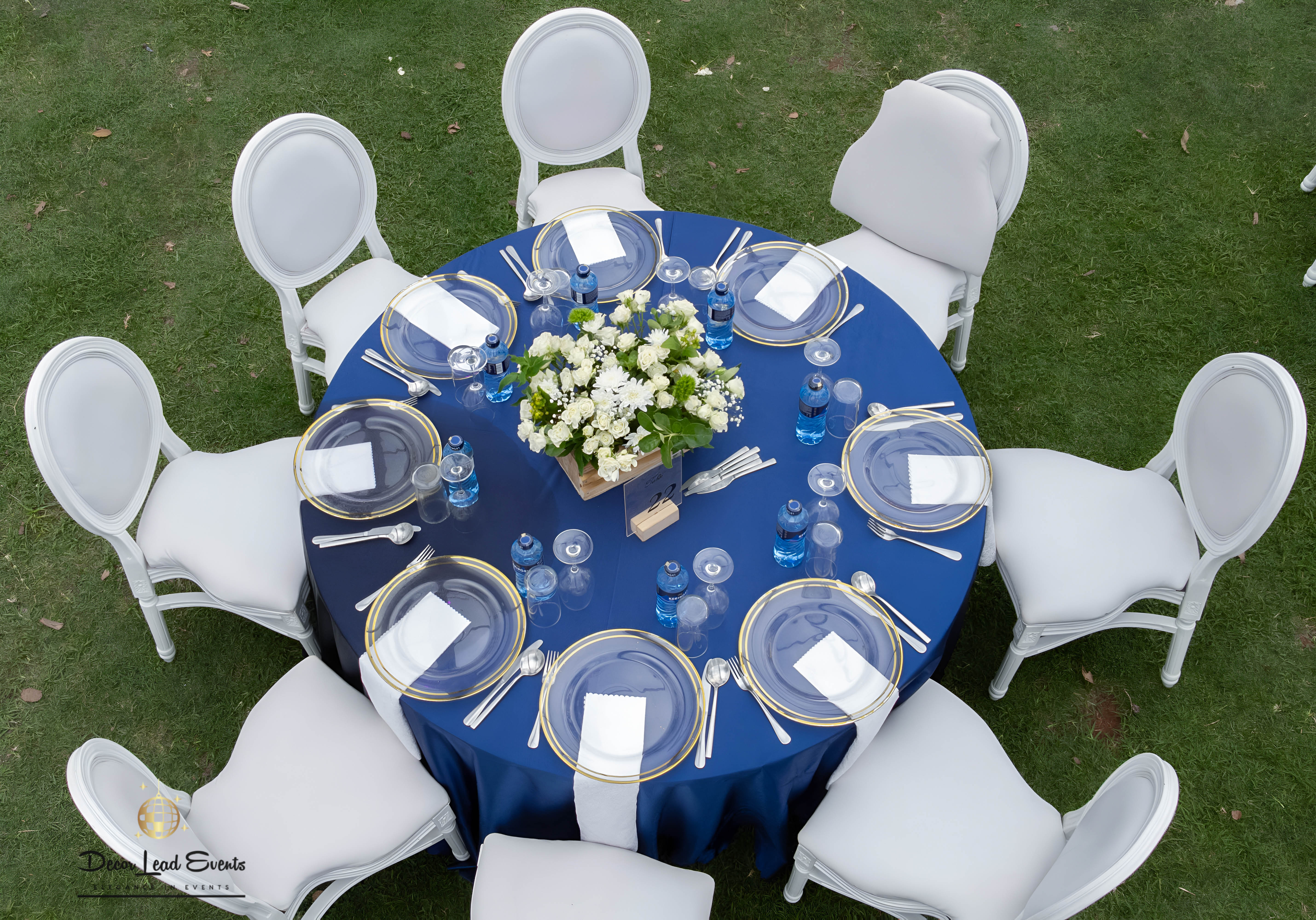 Aerial view of a round table set up with a navy blue tablecloth, gold-rimmed clear glass chargers, white napkins, wine glasses, and a white floral centerpiece.