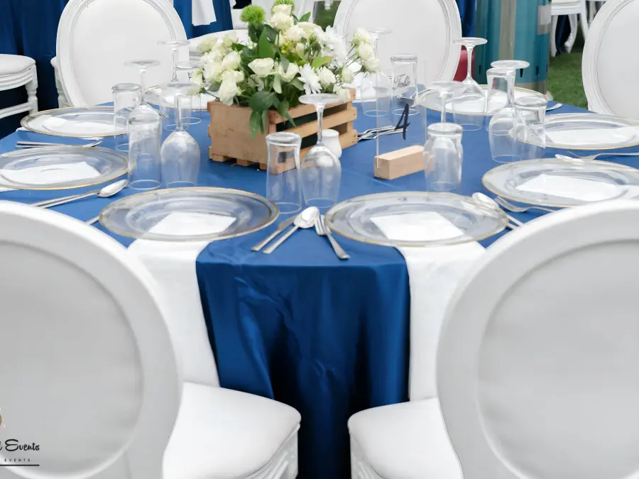 Medium shot of a round table setup with navy blue tablecloth and white round-back chairs, featuring a wooden crate centerpiece.