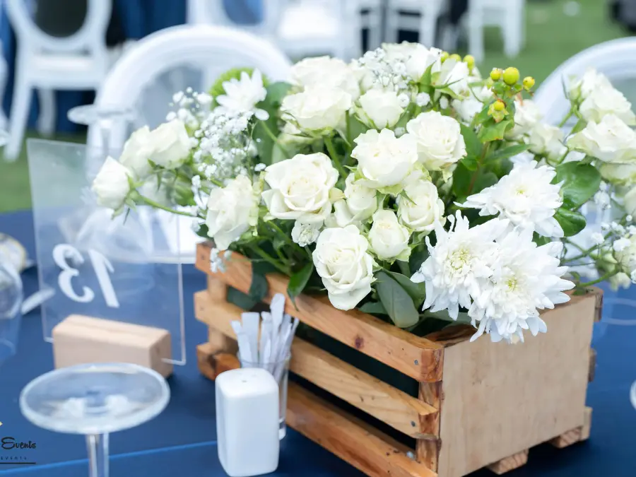 Close-up of a white rose floral arrangement in a rustic wooden crate on a blue tablecloth, with table number 13 visible.