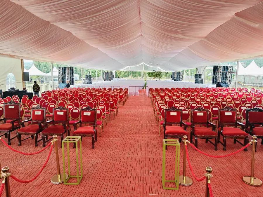 Large white tent interior set up for a formal event or funeral, featuring a red carpet aisle and rows of red banquet chairs.