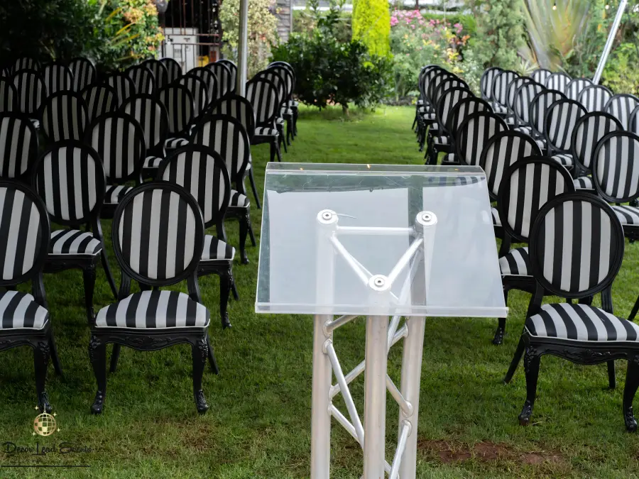 Outdoor ceremony setup on a lawn with rows of black and white striped oval-back chairs and a clear acrylic podium.