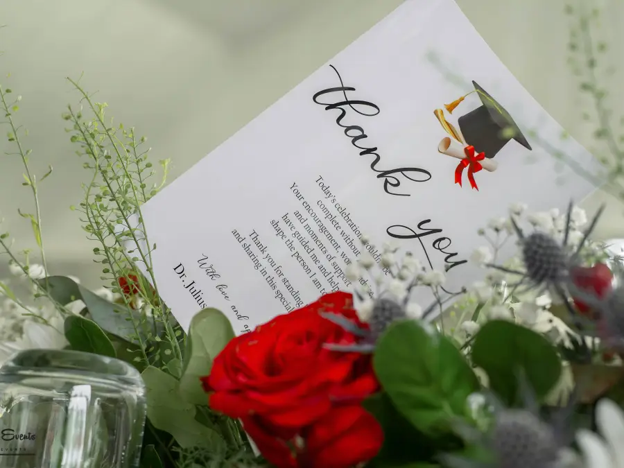 Close-up detail of a "Thank You" card with a graduation cap icon tucked into a floral centerpiece of red roses and white fillers.