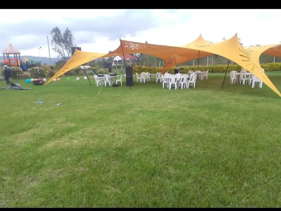 Yellow stretch tent set up on a lawn providing shade for white plastic chairs and tables, with a playground in the background.