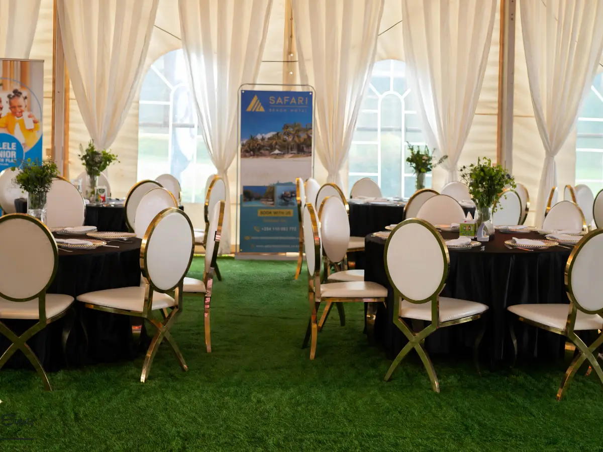 Elegant tent interior with black round tables, white gold-leg chairs, and white draped walls, with a Safari Park Hotel banner visible.