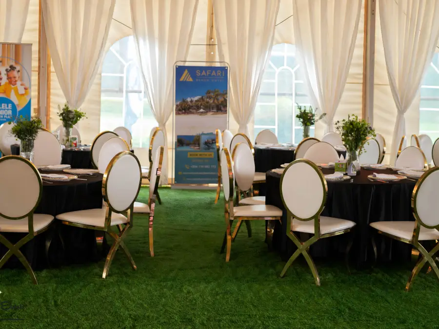 Elegant tent interior with black round tables, white gold-leg chairs, and white draped walls, with a Safari Park Hotel banner visible.