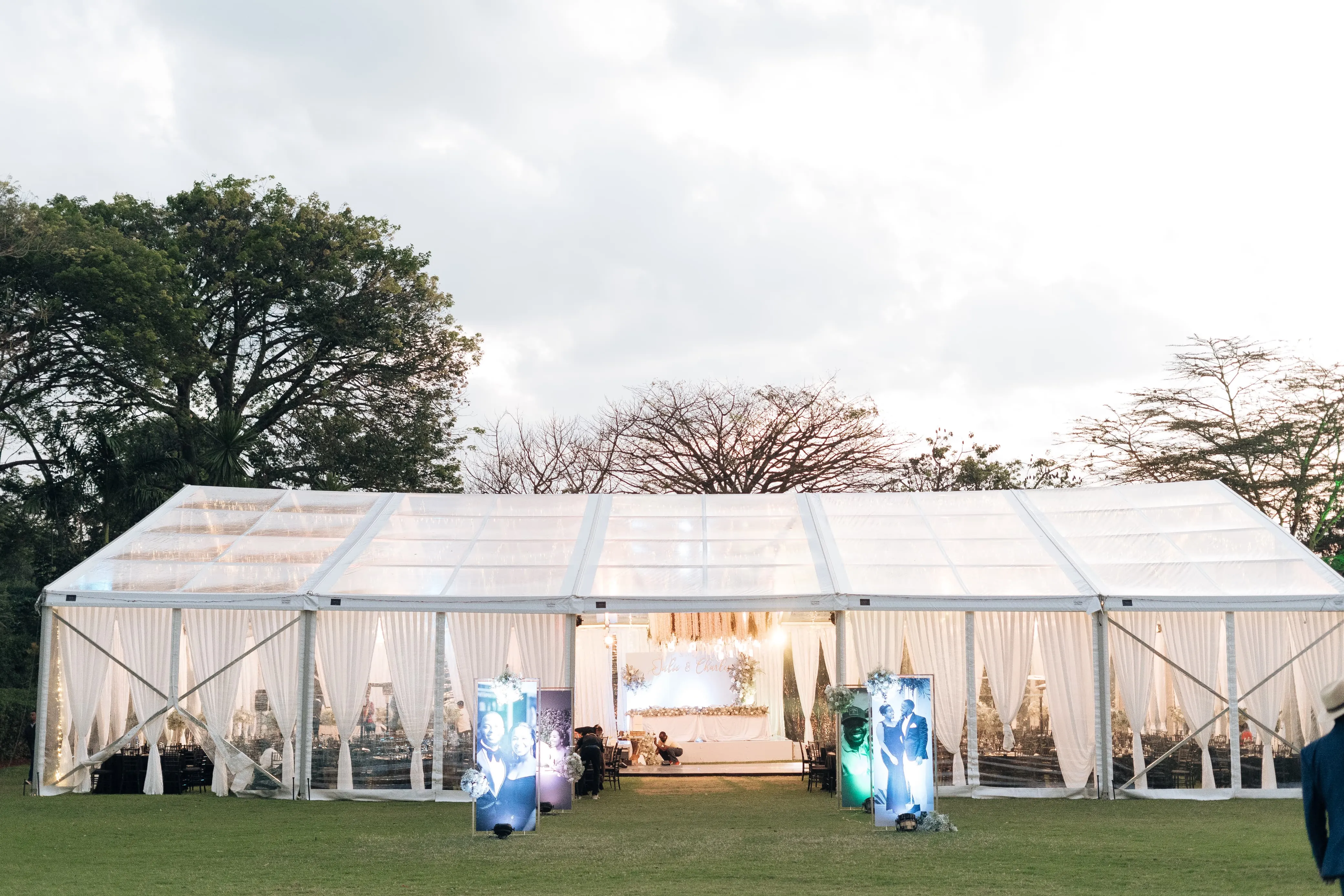 Exterior evening view of a large clear-span marquee tent with white drapes and warm lighting, set against a tree-lined backdrop.