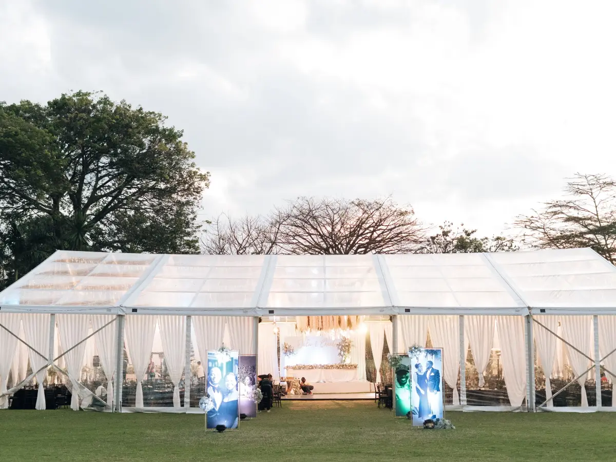 Exterior evening view of a large clear-span marquee tent with white drapes and warm lighting, set against a tree-lined backdrop.
