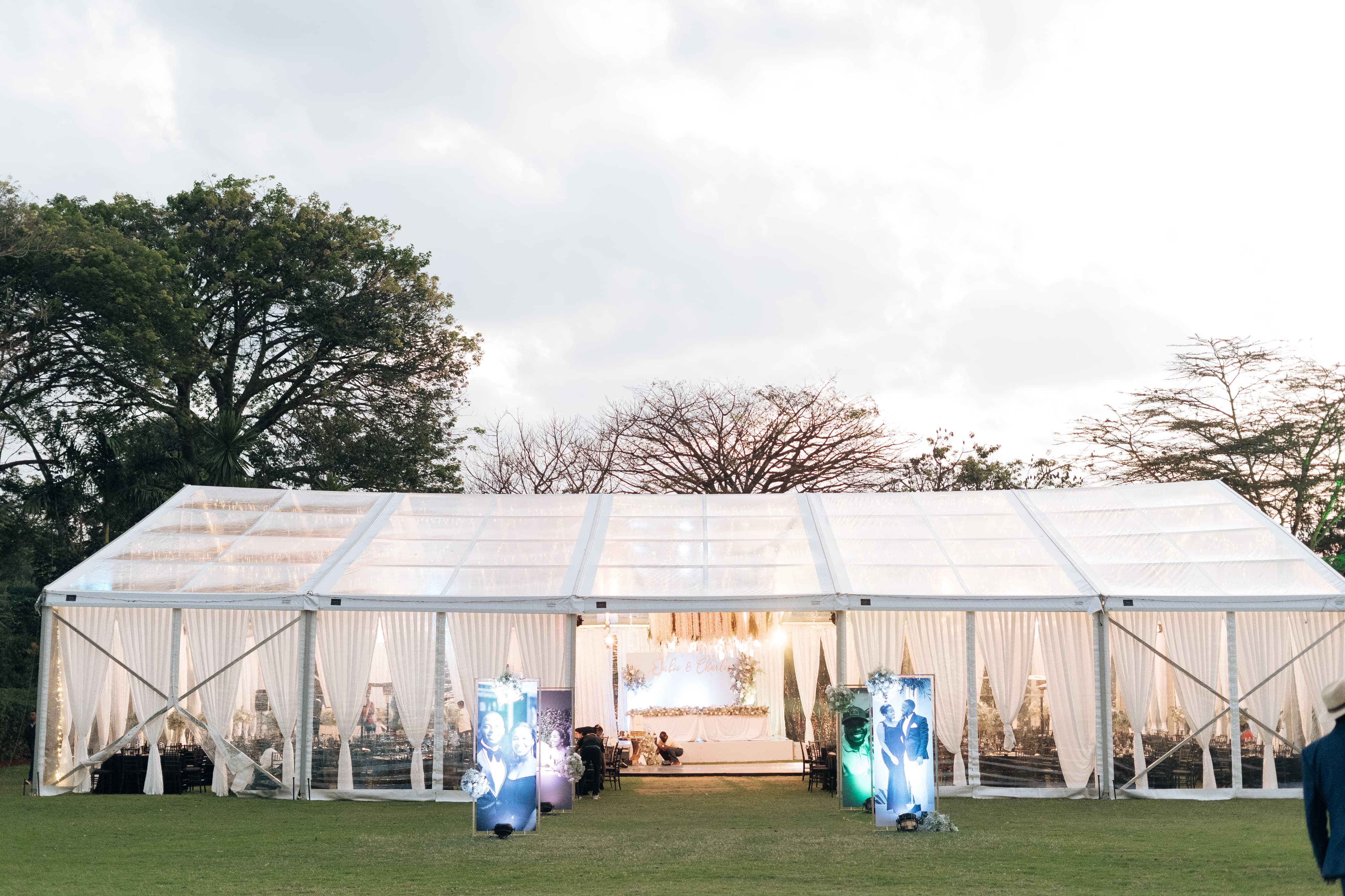 Exterior evening view of a large clear-span marquee tent with white drapes and warm lighting, set against a tree-lined backdrop.