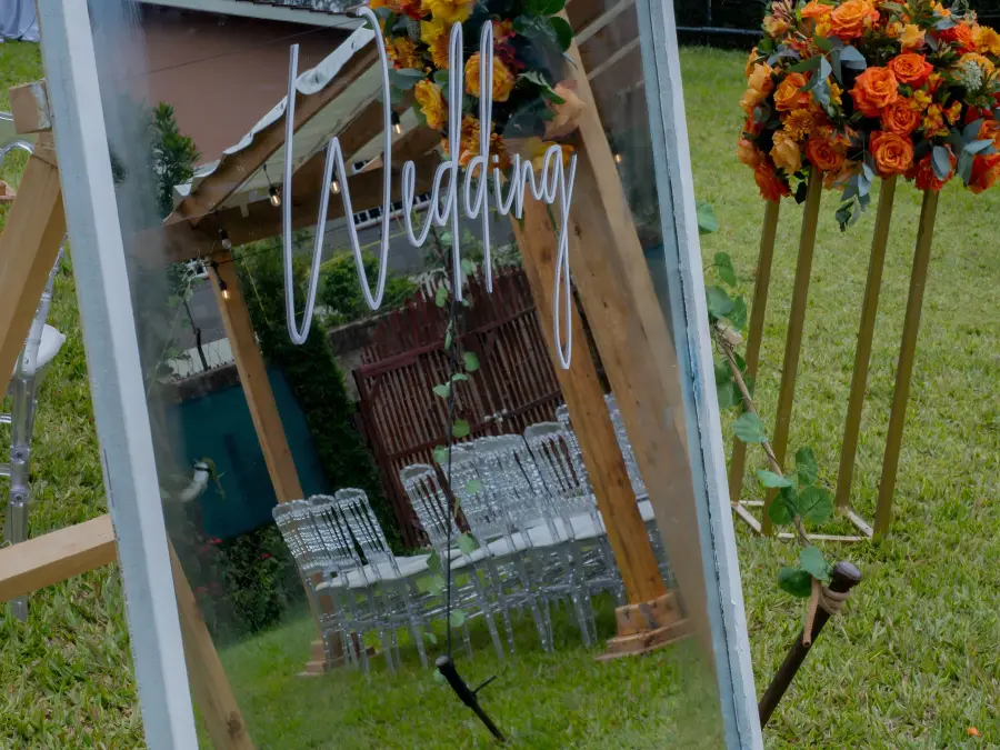 Ornate floor-standing mirror welcome sign for "Sonia & Ombaka" wedding, decorated with a garland of orange and yellow roses.