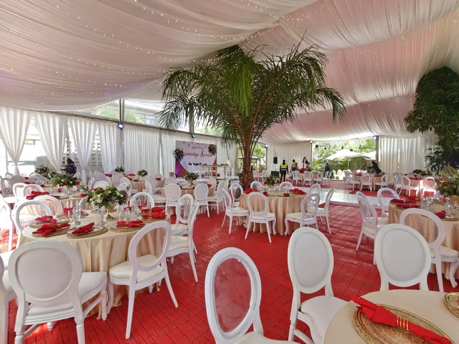 Interior of a white tent with a red tiled floor, beige round tables, white Louis chairs, and a large live palm tree integrated into the space.