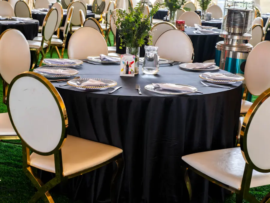 Round table setting with black tablecloth, silver tableware, and white oval-back chairs with gold legs, next to a patio heater.