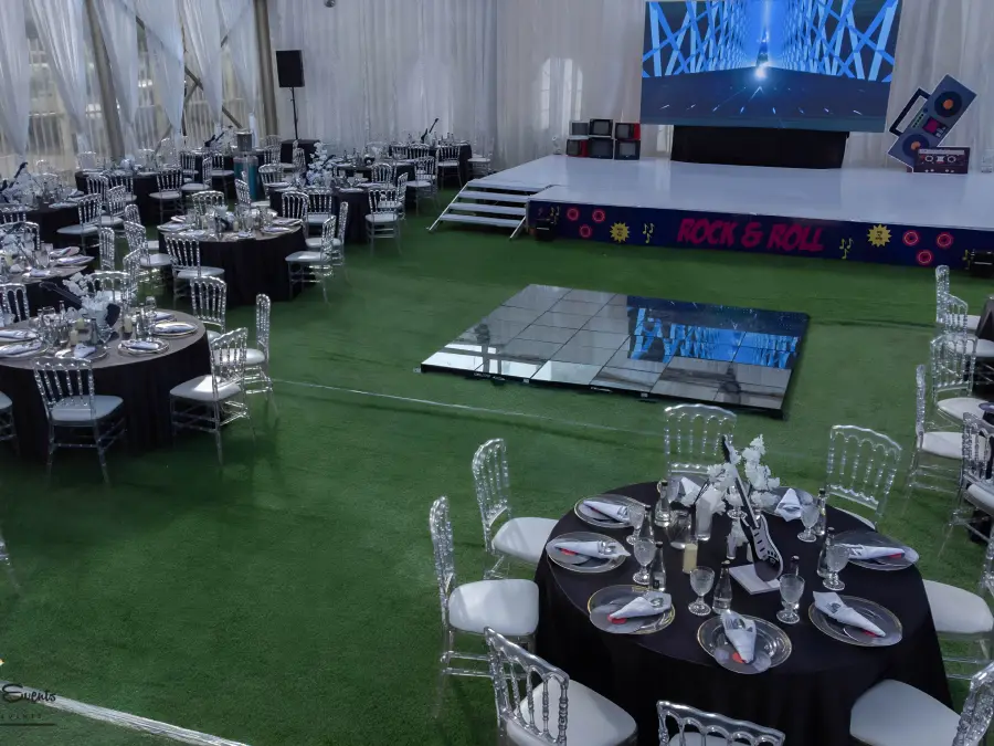 Spacious event hall setup with a "Rock & Roll" theme, featuring a large stage screen, black tables, and silver Chiavari chairs.