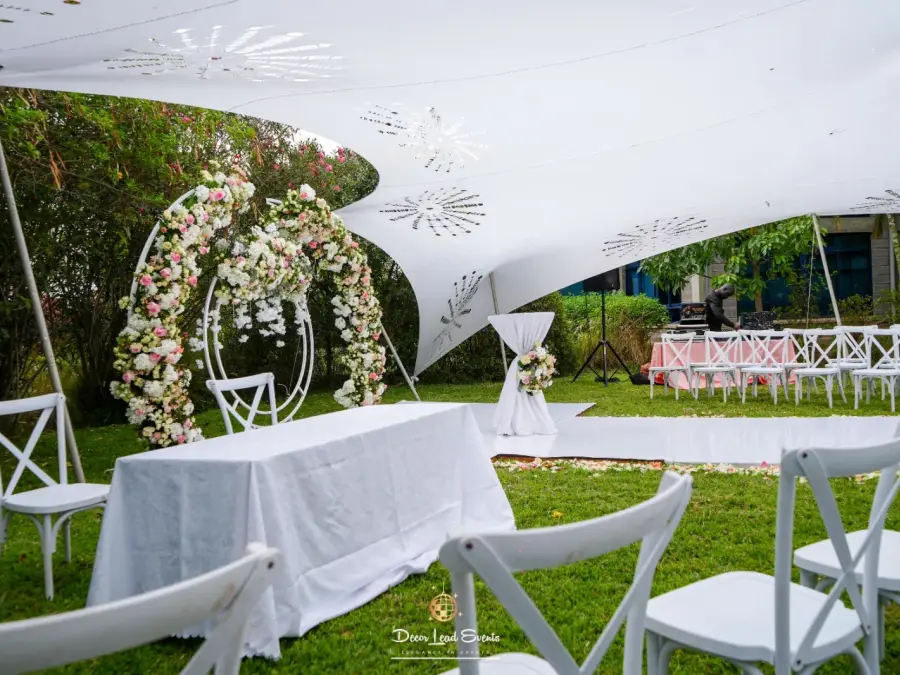 Outdoor wedding ceremony setup under a white cut-out stretch tent, featuring white cross-back chairs and a floral arch altar.
