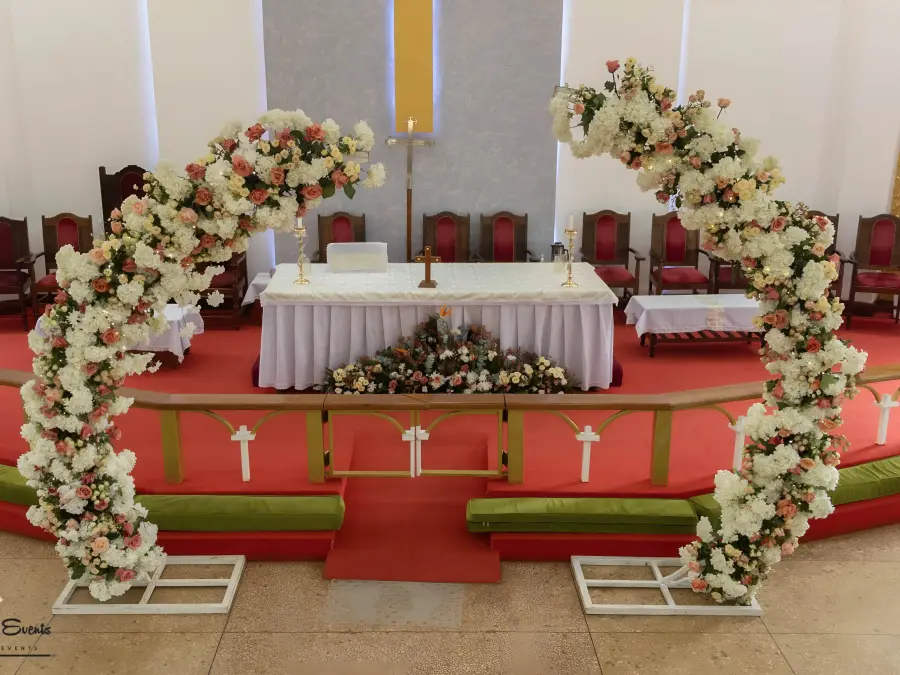 Church altar decorated with a split floral arch of white and peach roses, set on a red carpet.