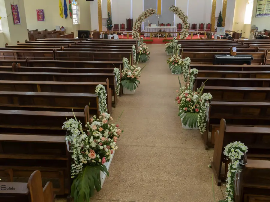 Wide view of a church aisle lined with wooden pews featuring floral arrangements, leading to a matching floral arch at the altar.
