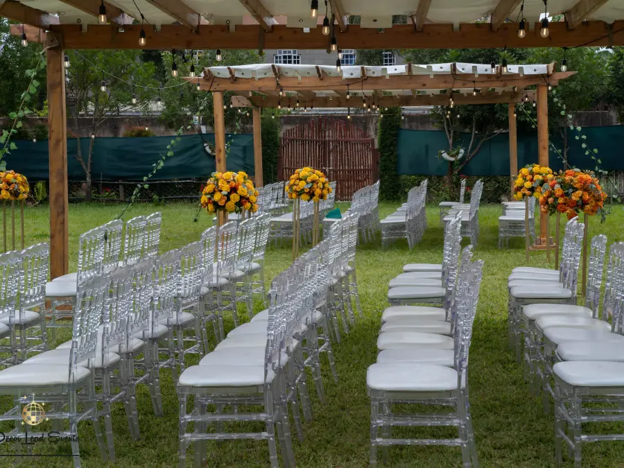 Outdoor wedding ceremony arrangement under a pergola with clear Chiavari chairs and vibrant orange and yellow floral pedestals.