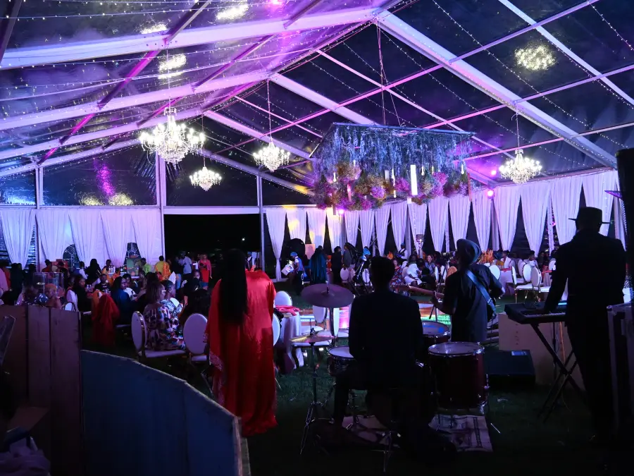 Nighttime interior of a clear-roof tent with a live band performing on stage and guests seated at tables under chandeliers.