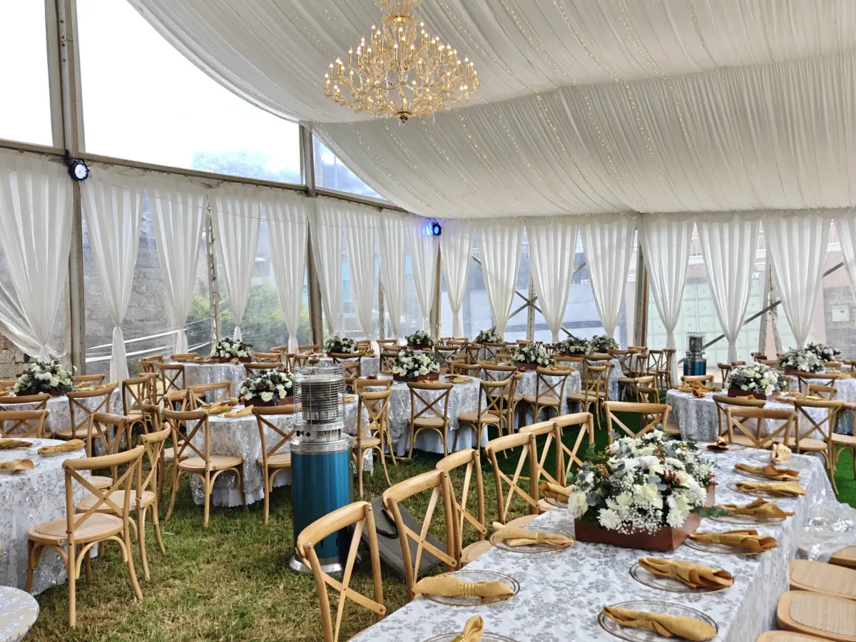Interior of a white marquee tent with draped ceilings, a crystal chandelier, and round tables with white linens and silver leaf patterned overlays.