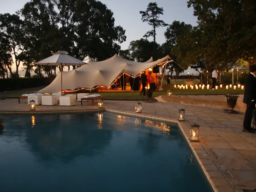 Evening shot of a large beige stretch tent set up near a pool, with warm string lighting and guests milling about.