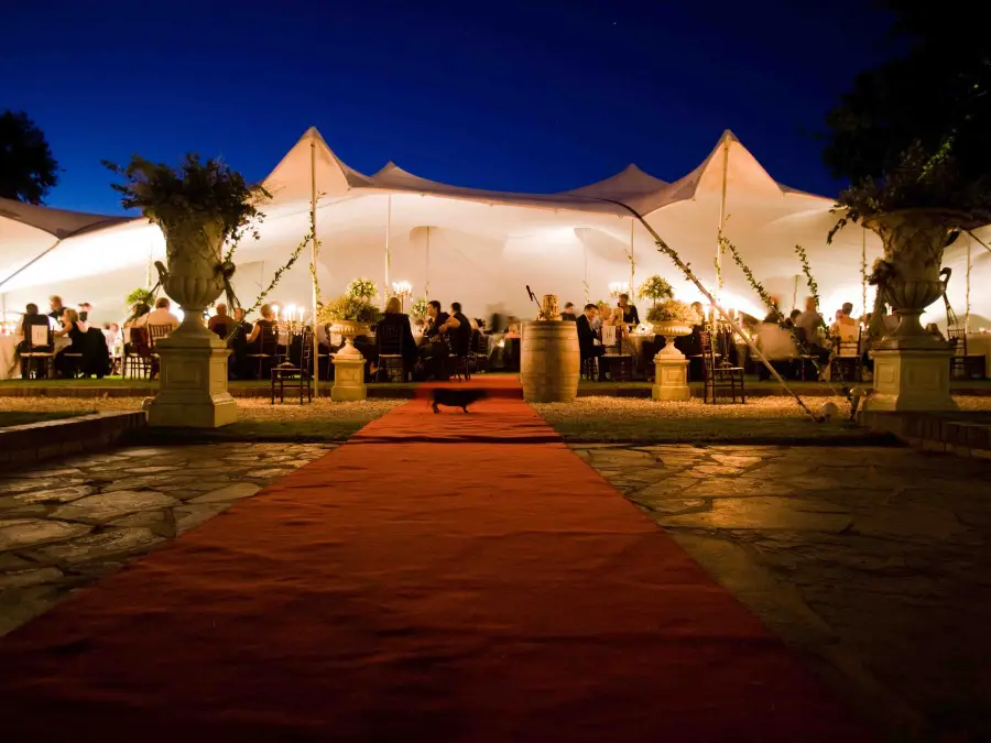 Nighttime view of a white stretch tent with a red carpet entrance, warm interior lighting, and guests dining at tables.
