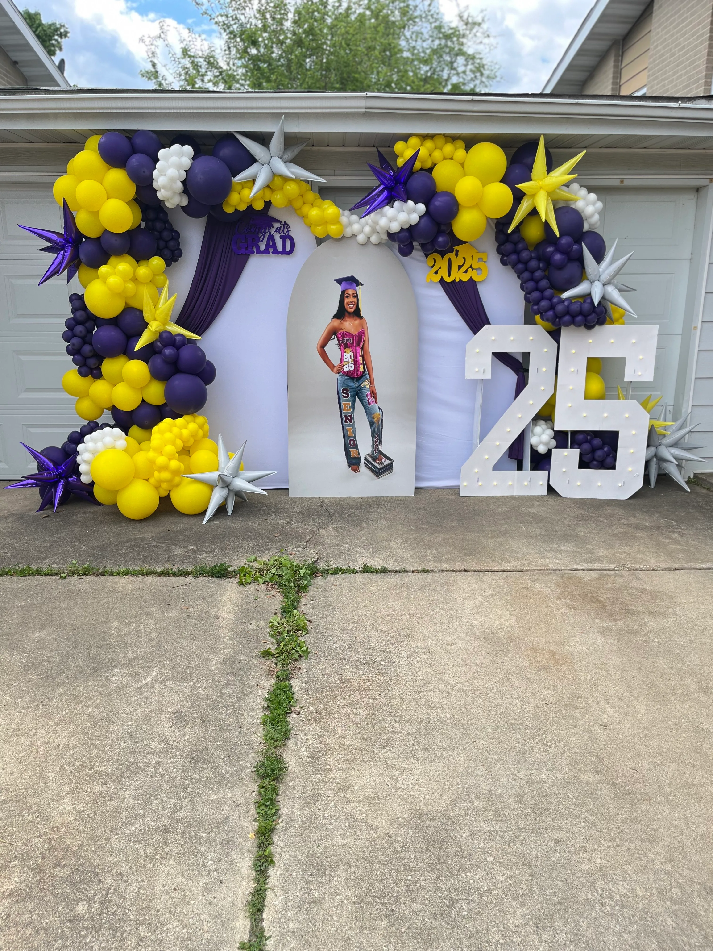 Graduation party display with balloon garland, fabric backdrop, life size graduate cutout, and lit marquee numbers.